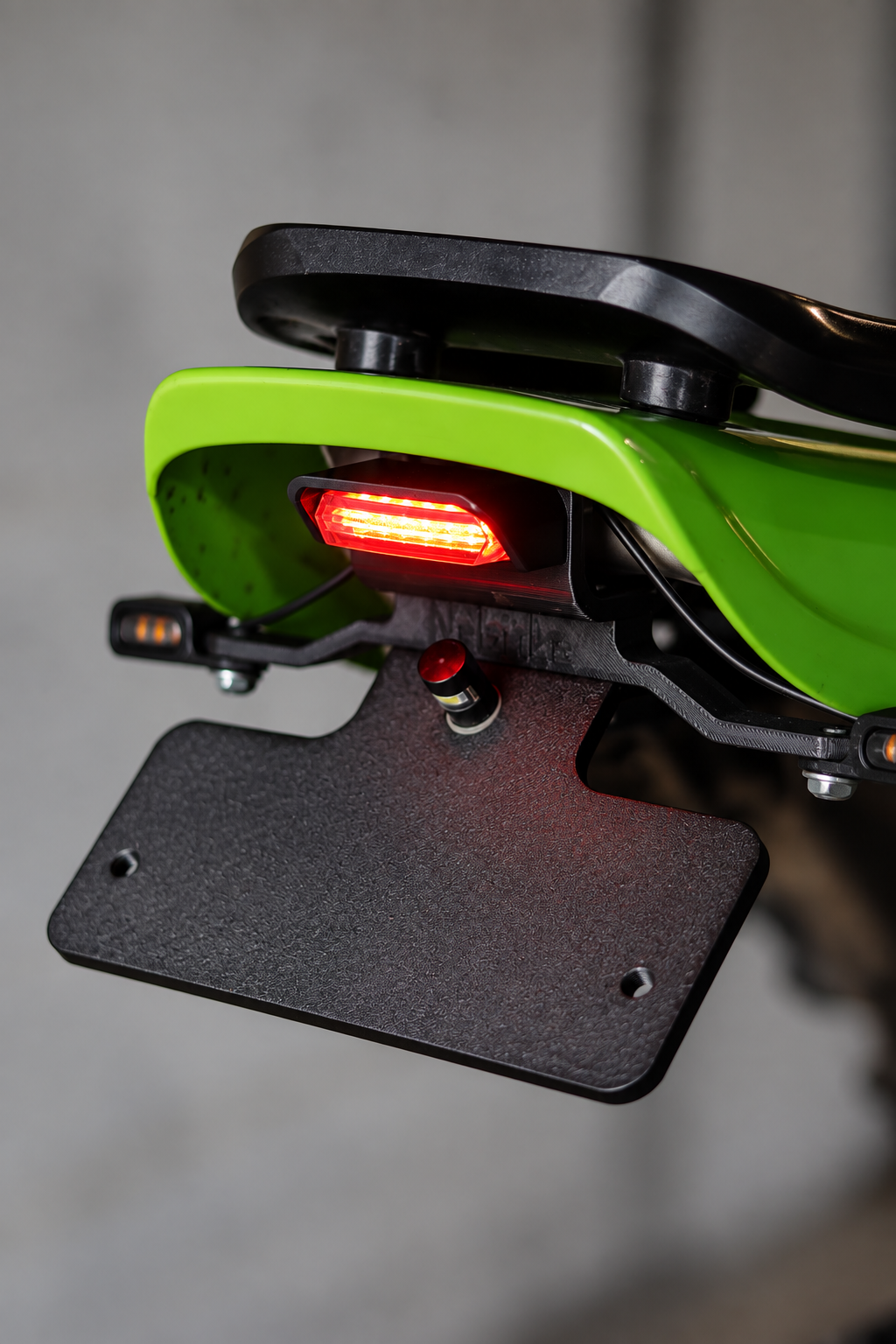 Close-up of a bright green and black motorcycle or scooter rear rack with a red illuminated taillight and a black license plate frame, shown against a blurred gray background.